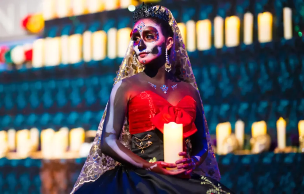 a woman dressed for dia de los muertos complete with head dress, calavera makeup, holding a prayer candle looking to the left. Prayer candles in rows in the background