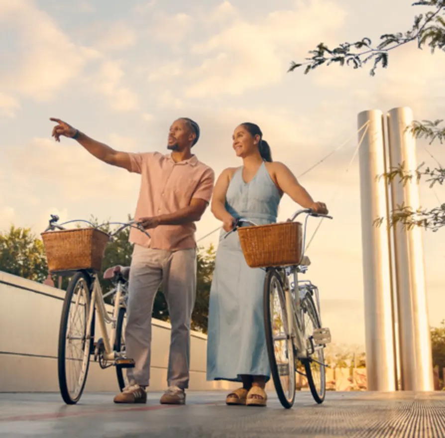 two people standing while holding their bikes. Man on the left pointing to something off in the distance to the left as woman looks in the same direction