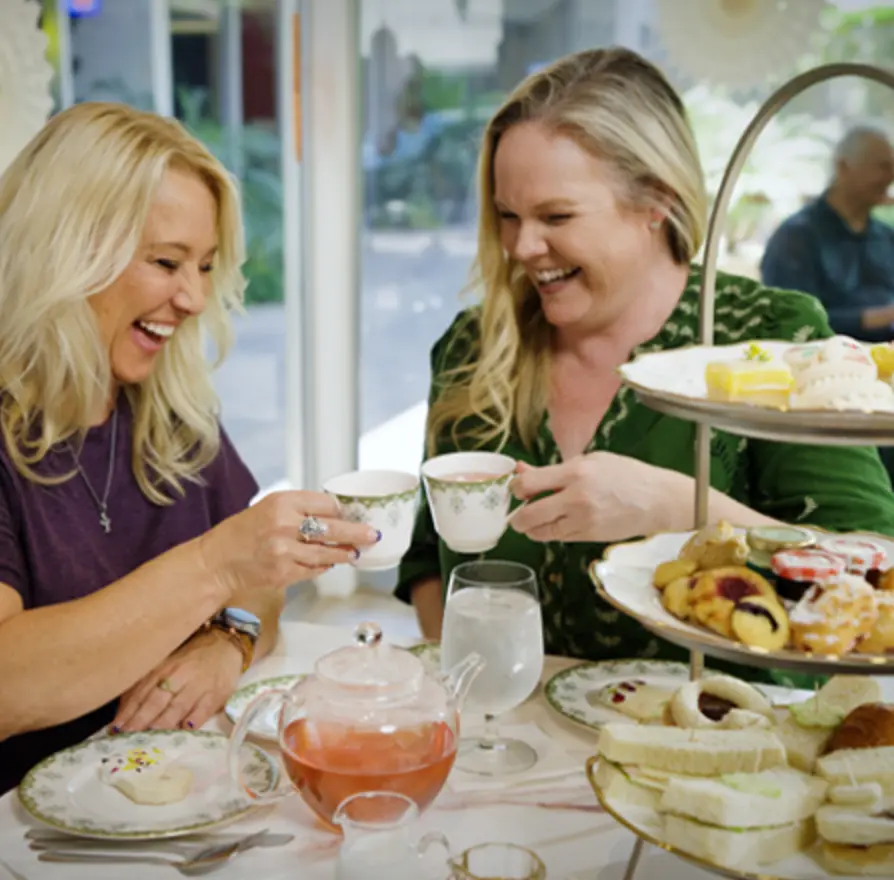 two women having afternoon tea in a tea room, laughing and toasting their tea cups. On the table are an assortment of scones, sandwiches and sweets.