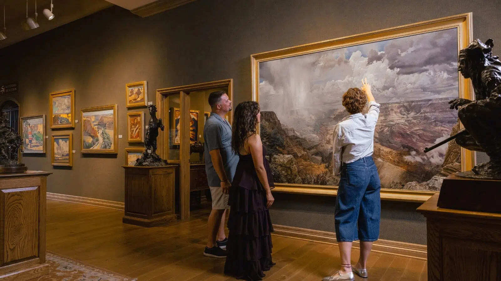 People point toward a large landscape painting of the Grand Canyon in an art gallery, with bronze sculptures displayed nearby.