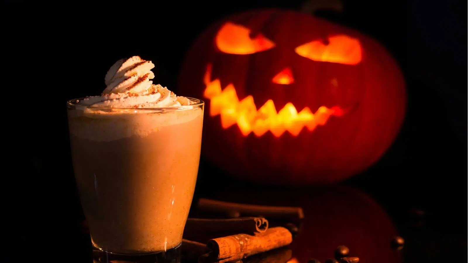 A creamy spiced drink topped with whipped cream next to cinnamon sticks, with a glowing jack-o'-lantern in the dark background.